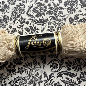May include: A skein of cream-colored thread wrapped in a black and gold paper label. The label features the text Art. 65, Lilly, and Daisy, along with a circular icon depicting a flower. The thread is textured and wound in a bundle.