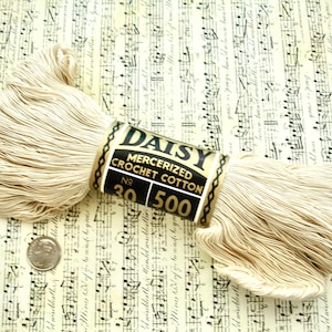 May include: A spool of Daisy mercerized crochet cotton in a light beige color, with the label visible. The label is black and gold with the text "Daisy Mercerized Crochet Cotton No. 30 500". A US dime is next to the spool for size comparison. The background is sheet music.