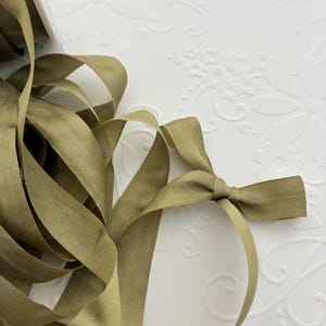 May include: Close-up of olive green ribbon with a small bow tied at the end. The ribbon is lying on a white surface with a floral pattern. The ribbon appears to be made of a soft, woven material.