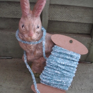 May include: A brown rabbit figurine with pink ears and a blue garland around its neck. A spool of light blue, sparkly tinsel is next to the rabbit. The tinsel is wrapped around a brown cardboard spool.