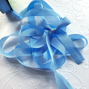 May include: A spool of light blue ribbon with a tied bow. The ribbon is arranged on a white surface with a floral embossed pattern. The ribbon's color is a soft, pastel blue.