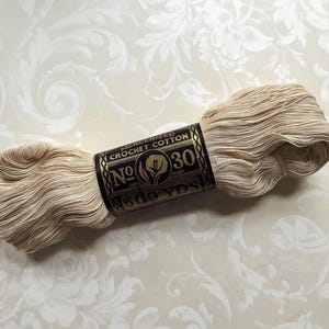 May include: A skein of cream-colored mercerized crochet cotton thread wrapped in a black paper label. The label features gold text reading No 30, Mercerized Crochet Cotton, and 500 YDS, along with a central circular logo graphic.