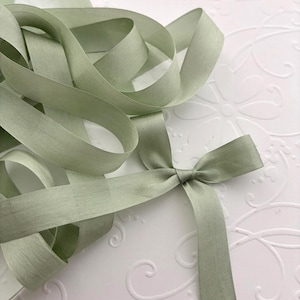 May include: Sage green ribbon, looped and tied into a bow. The ribbon is made of a textured material and is displayed on a white surface with a subtle floral pattern. The ribbon's color is a muted, earthy green.