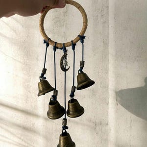 以下が含まれることがあります： 手作りのウィンドチャイム。ジュート織りのリング、ネイビーブルーのコード、ブロンズ色のベルが特徴です。三日月チャームが中央から吊り下げられています。チャイムには6つのベルがあり、屋内または屋外での使用を目的としています。