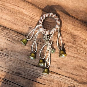 以下が含まれることがあります： 白い紐に小さな真鍮のベルが吊り下げられた、織りのリースが特徴の素朴なウィンドチャイム。装飾的な鍵と小さな花のチャームも付いています。ウィンドチャイムは木製の表面に表示されています。