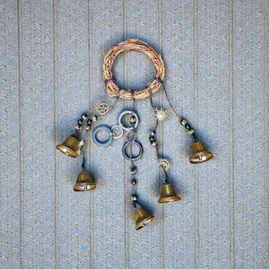 May include: A handcrafted wall hanging featuring a woven wreath with hanging brass bells and decorative charms. The design includes a pentagram, sun, and other celestial-themed accents, creating a rustic and bohemian aesthetic.