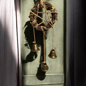 May include: A rustic wreath made from woven twigs, featuring a jute star and small green leaves. Three bronze bells are suspended below on jute twine. The wreath hangs against a green backdrop.