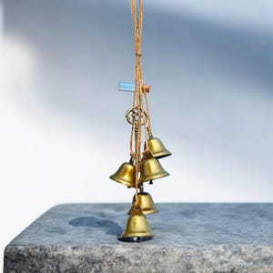 Può includere: Un ornamento sospeso con sei piccole campane color oro, sospese a un cordino color naturale. Sono attaccate anche una chiave decorativa e una piccola fiala di vetro. Le campane sono disposte in un gruppo, creando un'estetica delicata.