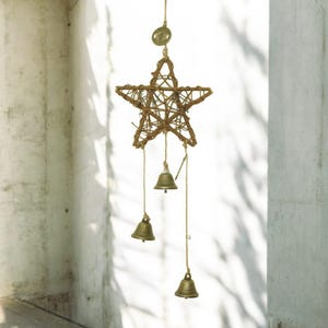 Könnte beinhalten: Ein sternförmiger Windspiel aus geflochtenen braunen Zweigen. Drei kleine Messingglocken hängen darunter. Das Windspiel hängt an einer Schnur, mit einem kleinen kreisförmigen Anhänger oben.