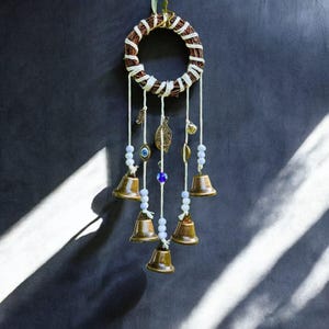Puede incluir: Un colgante de pared artesanal con un anillo tejido marrón y detalles de cuerda blanca. Cuenta con campanas de bronce, cuentas, dijes de hojas de metal y un amuleto de ojo turco, suspendidos por cuerdas blancas.