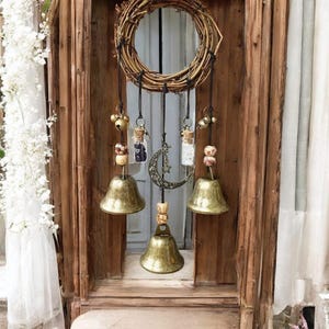 May include: A rustic wind chime with a woven wreath at the top. Below hang three brass bells, glass vials with cork stoppers, and a crescent moon charm. The wind chime is suspended against a wooden door.