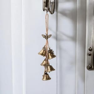 May include: A decorative wind chime featuring five small, antique brass bells suspended from a natural twine cord. A brass butterfly and decorative charms accent the bells. The chime hangs from a silver door handle against a white door.