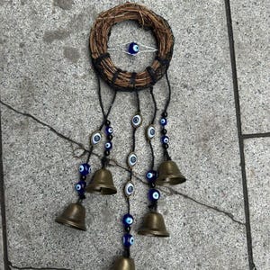 以下が含まれることがあります： 織り込まれたリースと中央の青い目のチャームが特徴の装飾的な壁掛け。黒いコードは真鍮のベルと青と白の邪視ビーズのストリングを吊り下げており、ユニークで人目を引くデザインを作り出しています。