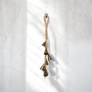 Peut inclure: Suspension décorative composée de petites cloches de couleur bronze suspendues à une corde naturelle. Les cloches sont coniques et de tailles légèrement différentes. La corde est bouclée sur une tête de clou en métal, créant une esthétique rustique et bohème.