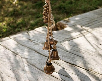 Puede incluir: Un carillón de viento de bronce con pequeñas campanas y cuentas decorativas. El carillón está suspendido de una cuerda natural y presenta un dije de media luna. El fondo muestra una terraza de madera y follaje verde.