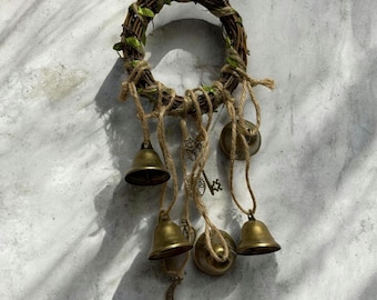 Puede incluir: Adorno de pared circular rústico con una base de ramitas tejidas adornada con vegetación artificial. Varias campanas de latón de varios tamaños cuelgan de hilo de yute, junto con una llave pequeña y un dije de media luna.
