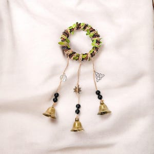 May include: A decorative wreath with a woven brown and green circular base, decorated with green leaves. Three strands hang down, each with black beads and a brass bell. Charms include a sunburst and a Celtic knot.