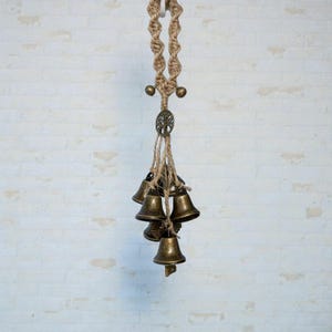 May include: A hanging decorative piece featuring a macrame rope design with small brass bells. The bells are suspended from a natural-colored rope, with a metal tree of life charm. The bells are various sizes and shapes.