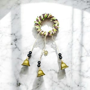 May include: A circular wall hanging with a woven brown base, decorated with green leaves. Three strands hang down, each with a brass bell, black beads, and silver charms. The charms include a sun, a tree of life, and a Celtic knot.