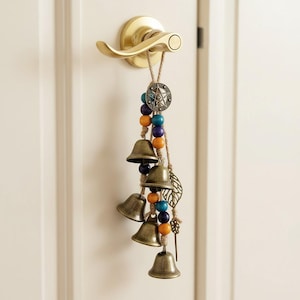 May include: A door chime featuring bronze bells, colourful wooden beads, and a starburst pendant. The chime is suspended from a natural fibre cord attached to a gold door handle. A leaf and key charm hang below the bells.
