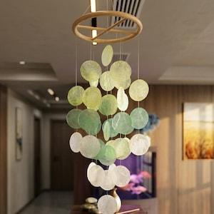May include: A hanging mobile featuring a wooden ring and strings of iridescent, circular shell pieces in shades of green and white. The mobile is suspended in a well-lit room, creating a decorative and airy ambiance.