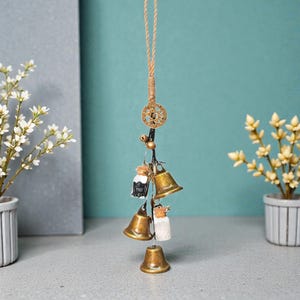 Puede incluir: Un carillón de viento colgante con pequeñas campanas de latón y elementos decorativos. El carillón incluye un dije en forma de estrella, pequeños frascos y una cuerda natural para colgar. Las campanas son de varios tamaños y formas.
