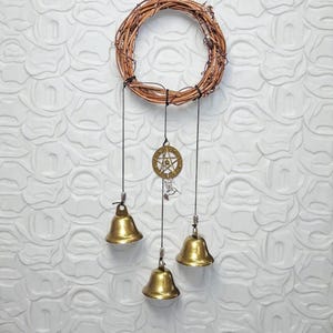 Könnte beinhalten: Ein handgefertigter Windspiel mit einem kreisförmigen geflochtenen Kranz. Drei Messingglocken hängen darunter, aufgehängt an schwarzen Schnüren. Ein dekorativer Messinganhänger mit Sternmuster ist ebenfalls am Kranz aufgehängt. Der Hintergrund ist eine strukturierte weiße Wand.