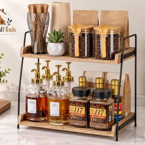 May include: A two-tier countertop organiser with a black metal frame and light wood shelves. The top shelf holds a glass, a small succulent, and two glass jars with bamboo lids and wooden spoons. The bottom shelf holds four glass pump bottles and two jars labelled The Blend 117.