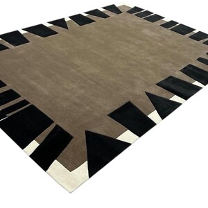 May include: A rectangular area rug featuring a solid taupe center field surrounded by a border of alternating black geometric shapes and white accents. The design consists of various black triangles and trapezoids arranged along the perimeter.