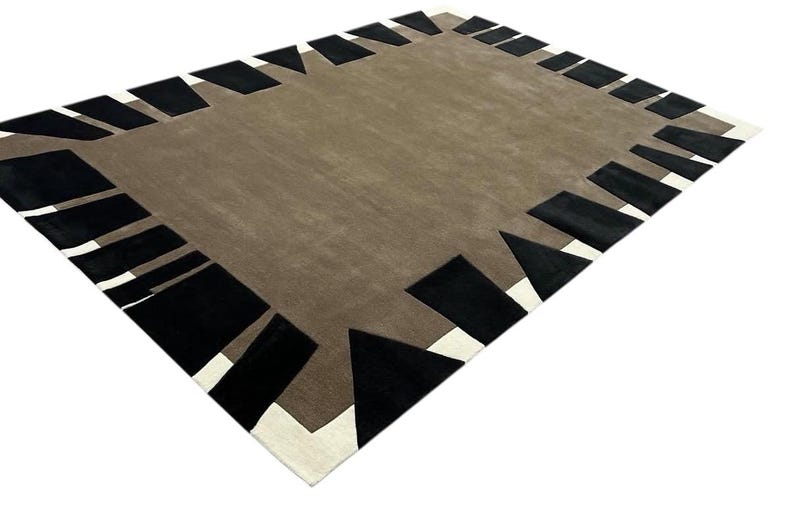 May include: A rectangular area rug featuring a solid taupe center field surrounded by a border of alternating black geometric shapes and white accents. The design consists of various black triangles and trapezoids arranged along the perimeter.