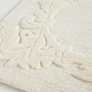 May include: An off-white area rug featuring a raised, tufted botanical pattern of leaves and vines along the border. The rug has a textured, dense pile construction with a monochromatic color scheme, showcasing a soft, neutral aesthetic throughout.