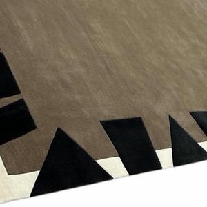 May include: A rectangular area rug featuring a large, solid taupe center field. The border consists of a white inner frame accented by a series of black geometric shapes, including triangles and rectangles, arranged along the outer edges.