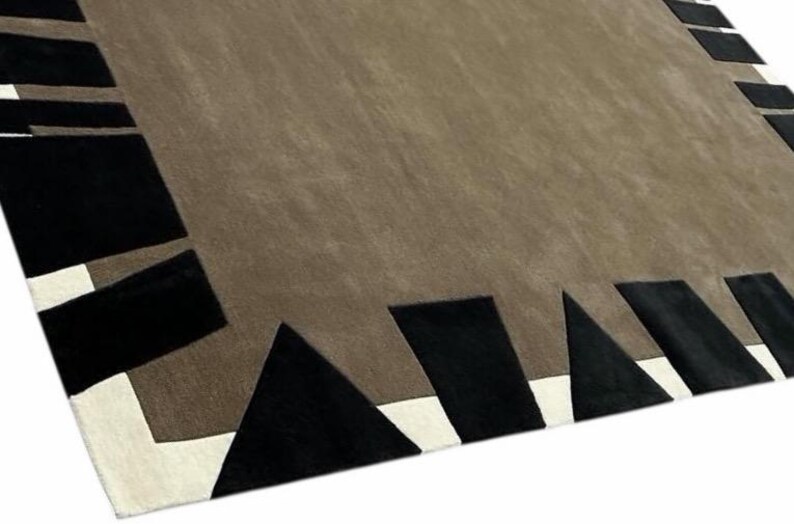 May include: A rectangular area rug featuring a large, solid taupe center field. The border consists of a white inner frame accented by a series of black geometric shapes, including triangles and rectangles, arranged along the outer edges.