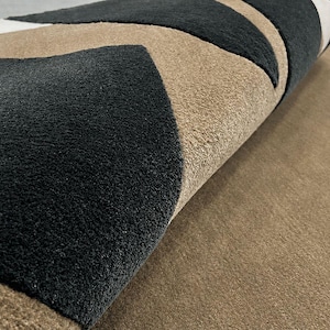 May include: A textured area rug featuring a geometric pattern of alternating black, beige, and white triangular shapes. The rug is partially folded, revealing the thick, plush pile construction and the distinct color-blocked design across the surface.
