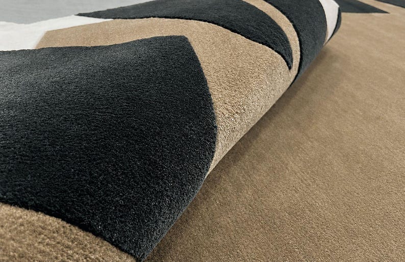 May include: A textured area rug featuring a geometric pattern of alternating black, beige, and white triangular shapes. The rug is partially folded, revealing the thick, plush pile construction and the distinct color-blocked design across the surface.