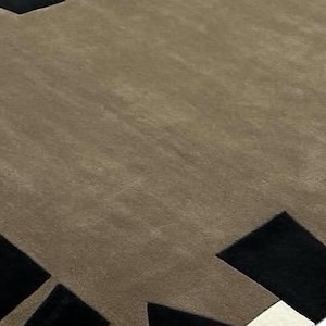 May include: A large area rug featuring a solid taupe center field surrounded by a geometric border. The border consists of alternating black rectangular and triangular shapes set against a thin white accent line along the outer edge.
