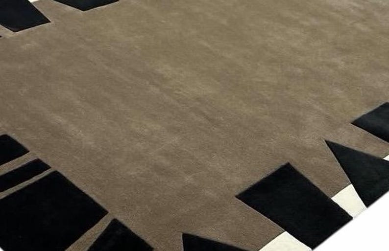 May include: A large area rug featuring a solid taupe center field surrounded by a geometric border. The border consists of alternating black rectangular and triangular shapes set against a thin white accent line along the outer edge.