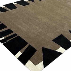 May include: A rectangular area rug featuring a solid taupe center field surrounded by a geometric border. The border consists of alternating black trapezoidal shapes and white triangular accents, creating a high-contrast frame around the rug.