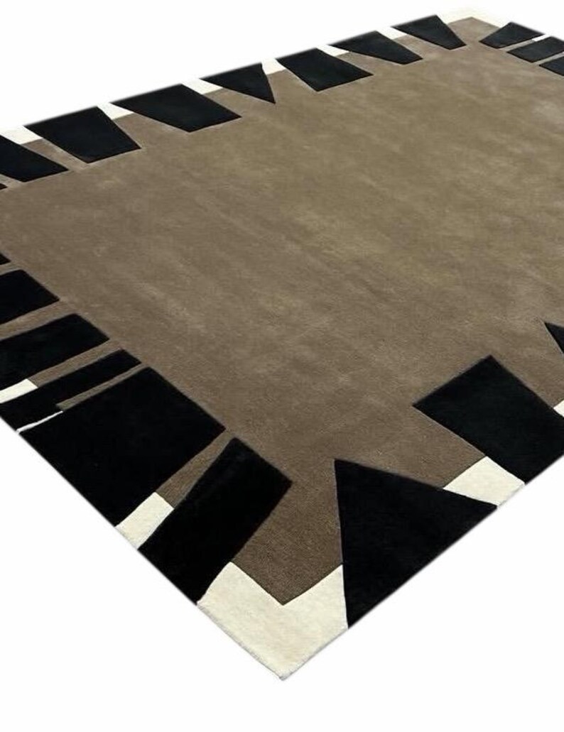 May include: A rectangular area rug featuring a solid taupe center field surrounded by a geometric border. The border consists of alternating black trapezoidal shapes and white triangular accents, creating a high-contrast frame around the rug.