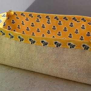 May include: A rectangular storage basket with a natural linen exterior and a bright yellow interior patterned with black floral motifs. The rim is trimmed with a yellow fabric featuring a black and white floral design. Gold-coloured buttons adorn the corners.