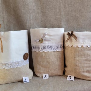 Op de afbeelding: Drie decoratieve jute zakken met witte bovenkanten en kanten of lint accenten. Elke tas heeft een ander versiering: een houten knoop, een goudkleurige broche en een bruine strik. De tassen zijn genummerd 1, 2 en 3.