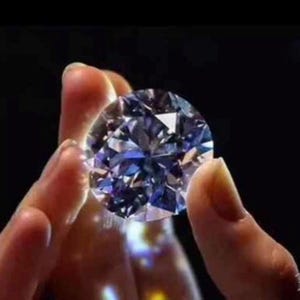 Könnte beinhalten: Ein großer, runder, facettierter Diamant mit Brillantschliff, der zwischen Fingern vor dunklem Hintergrund gehalten wird. Der Diamant reflektiert Blau- und Lilatöne und zeigt seine Klarheit und sein Funkeln.