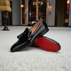 May include: Pair of black patent leather loafers with tassel detail. The shoes have a glossy finish and a red sole. The loafers are displayed on a white carpet.