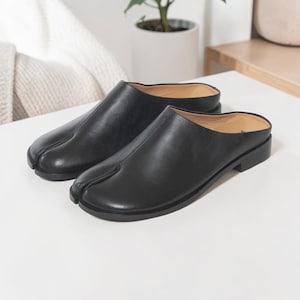 May include: A pair of black leather slip-on mules featuring a distinct split-toe design at the front. These shoes have a smooth finish, a low heel, and a tan-colored interior lining, showcasing a clean, minimalist silhouette for everyday wear.