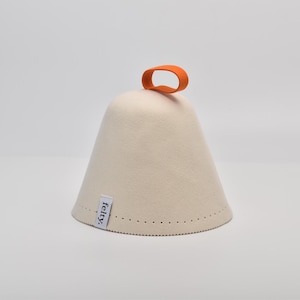 Puede incluir: Un gorro de sauna de fieltro color crema con un asa de bucle naranja. El gorro tiene forma cónica con la parte superior redondeada y un borde cosido. La palabra "felty." está impresa en una pequeña etiqueta blanca.