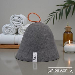 May include: A gray felt sauna hat with an orange loop handle. The hat has a small white tag that says "felty." Rolled white towels and a lit candle are in the background. The text "Ships Apr 15" is at the bottom.