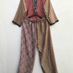 May include: A two-piece pajama set. The top is a long-sleeved shirt with a pink diamond pattern on the chest and tan sleeves with small brown dots. The pants are a mix of a floral pattern and a tan dotted pattern. The set is hanging on a wooden hanger.