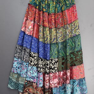 Könnte beinhalten: Ein langer Patchwork-Rock mit einer Vielzahl von bunten Paisley- und Blumenmustern. Der Rock besteht aus mehreren Stoffbahnen in Blau-, Rot-, Grün- und Schwarztönen. Er hängt an einem Holzbügel vor grauem Hintergrund.