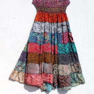 Könnte beinhalten: Ein farbenfrohes Patchwork-Kleid mit einem trägerlosen, gerafften Oberteil. Der Rock hat ein gestuftes Design mit verschiedenen gemusterten Stoffen in Rot-, Orange-, Blau-, Grün- und Lilatönen. Das Kleid hängt an einem Holzbügel vor weißem Hintergrund.