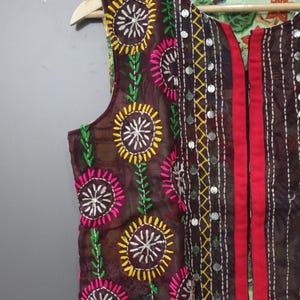 Puede incluir: Un chaleco marr&oacute;n con bordados florales coloridos. El chaleco presenta dise&ntilde;os de flores amarillas, rosas y blancas con tallos verdes. Tiene una raya roja en la parte delantera y est&aacute; decorado con peque&ntilde;os adornos plateados redondos.